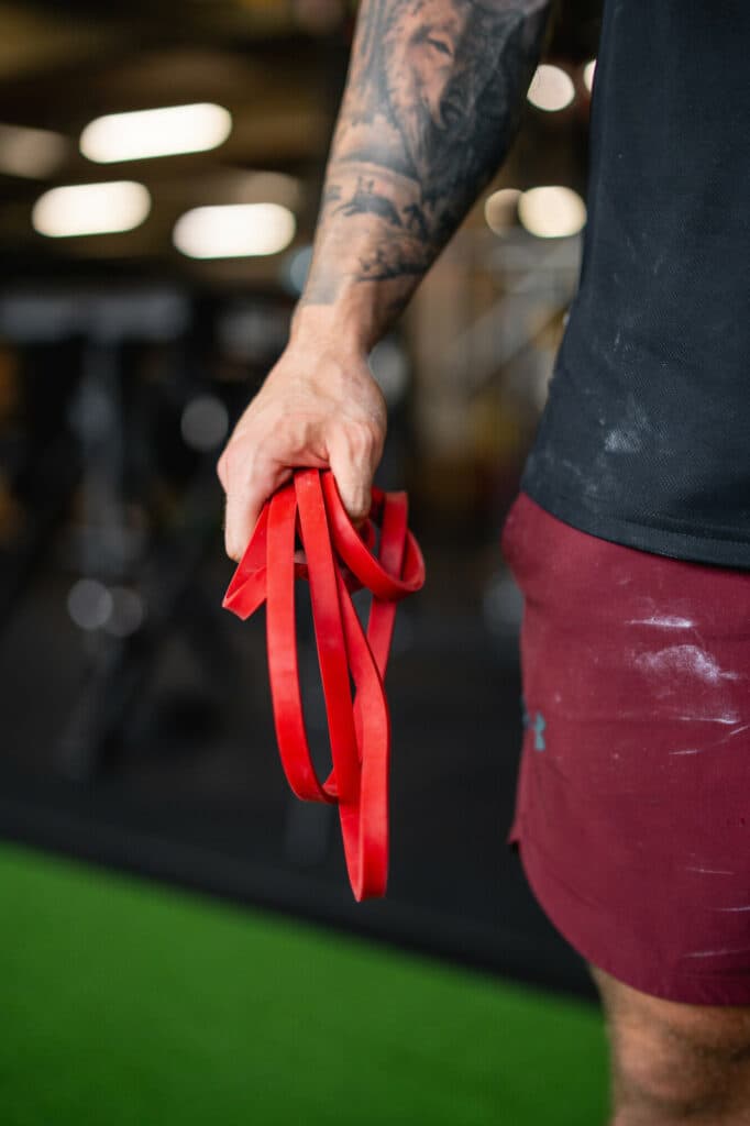 Eine tätowierte Hand hält ein rotes elastisches Widerstandsband vor einem unscharfen Fitnessstudio-Hintergrund.