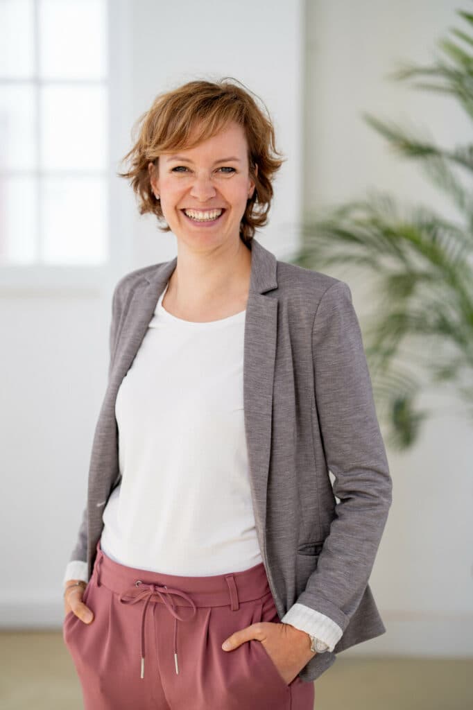 Eine lächelnde Frau mit kurzem braunem Haar trägt einen grauen Blazer über einem weißen T-Shirt und eine rosa Hose und steht vor einem hellen Hintergrund mit einer Pflanze.