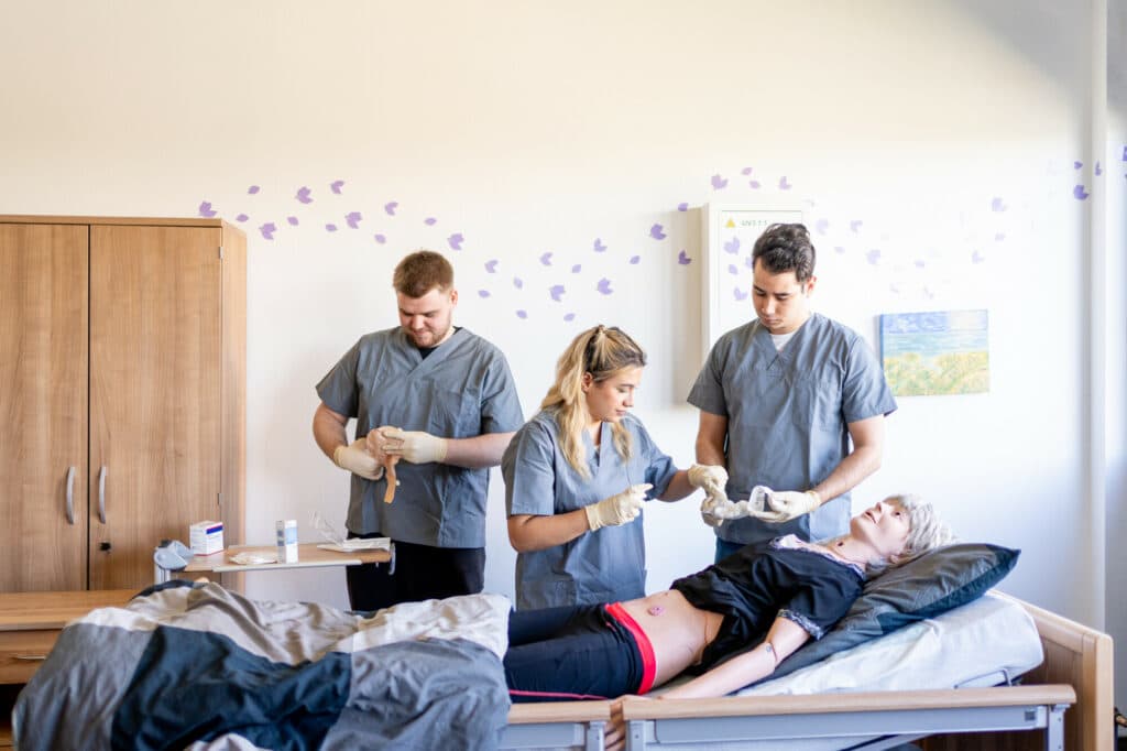Zwei Pfleger und eine Pflegerin in grauer Arbeitskleidung und mit Handschuhen versorgen eine liegende Patientin in einem hellen Krankenzimmer.