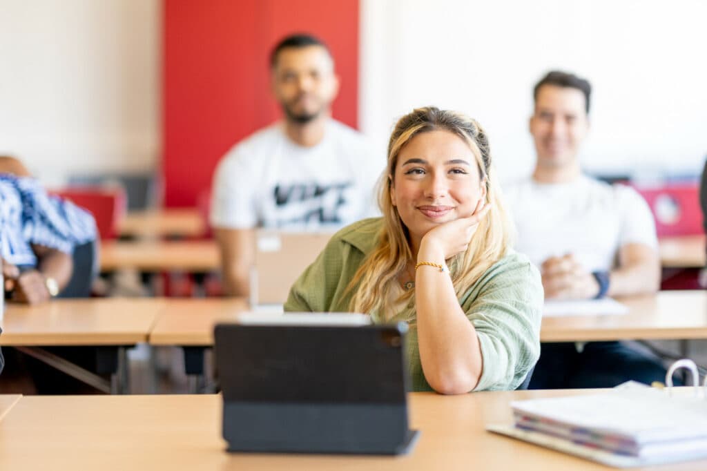 Eine junge Frau mit blonden Haaren sitzt an einem Tisch in einem Seminarraum und stützt ihr Kinn auf die Hand, während sie nach vorne blickt, im Hintergrund sitzen zwei weitere Personen an Tischen.