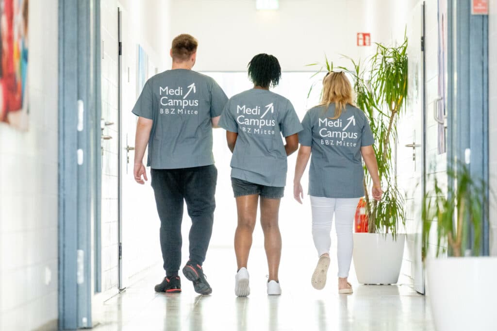 Drei Personen in grauen T-Shirts mit der Aufschrift Medi Campus BBZ Mitte gehen von der Kamera weg durch einen hellen Flur.