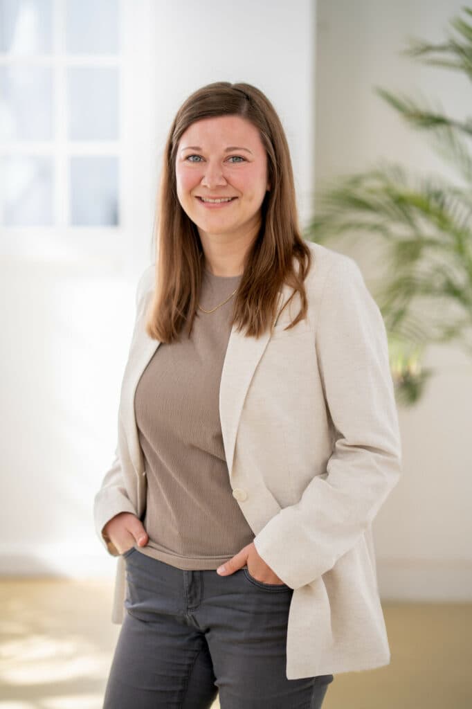 Eine lächelnde Frau mit braunen Haaren trägt einen hellen Blazer über einem braunen T-Shirt und steht in einem hellen Raum vor einer Pflanze.