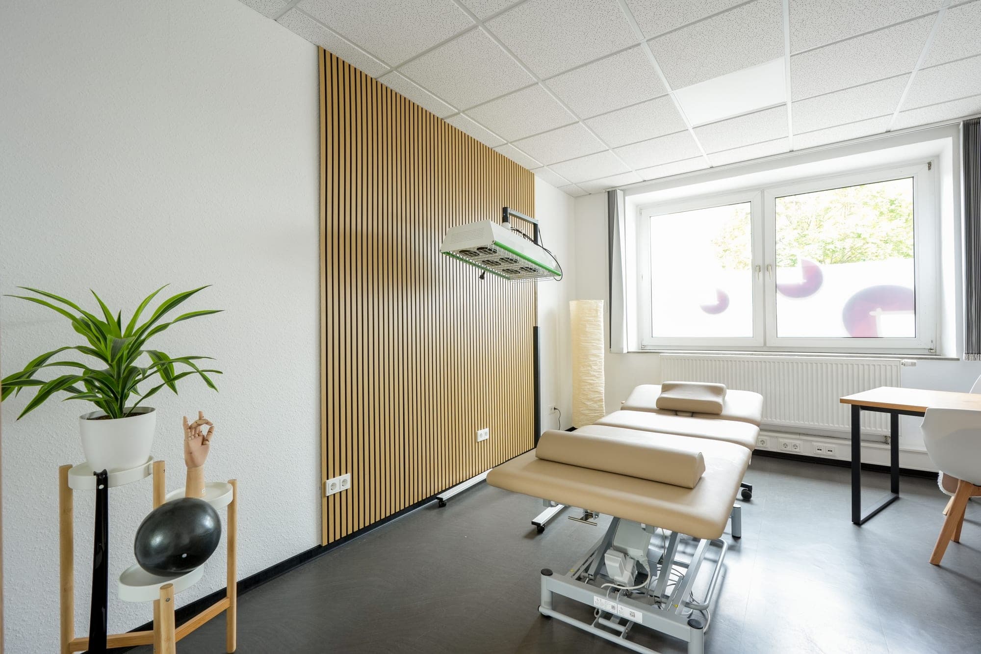 Ein heller Behandlungsraum mit einer beigen Massageliege, einer Wand mit vertikalen Holzlamellen, einer Zimmerpflanze auf einem Ständer und einem Fenster.