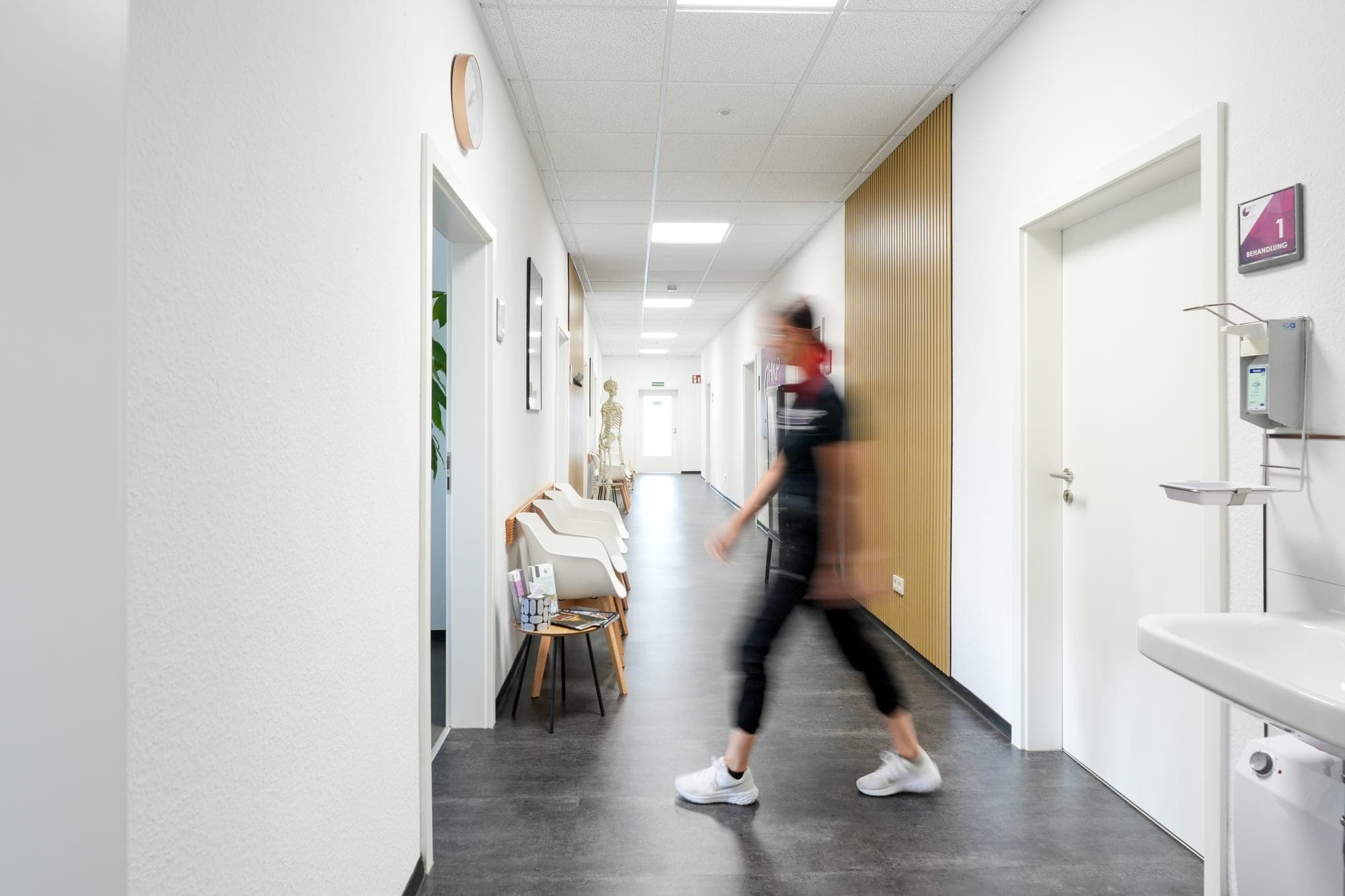 Eine verschwommene Person in Sportkleidung geht durch einen hellen, weißen Flur einer medizinischen Einrichtung.