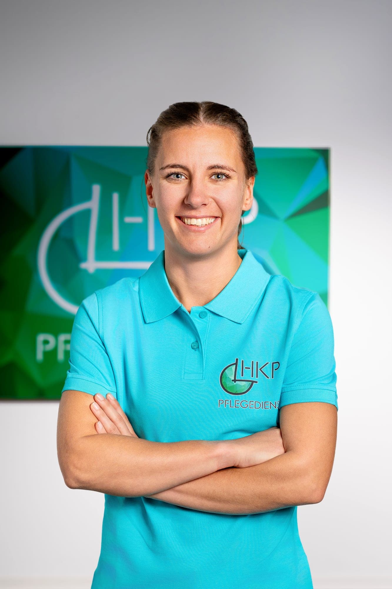 Eine lächelnde Frau mit braunen Haaren trägt ein türkises Poloshirt mit einem Firmenlogo und steht mit verschränkten Armen vor einem grünen, geometrisch gemusterten Hintergrund.