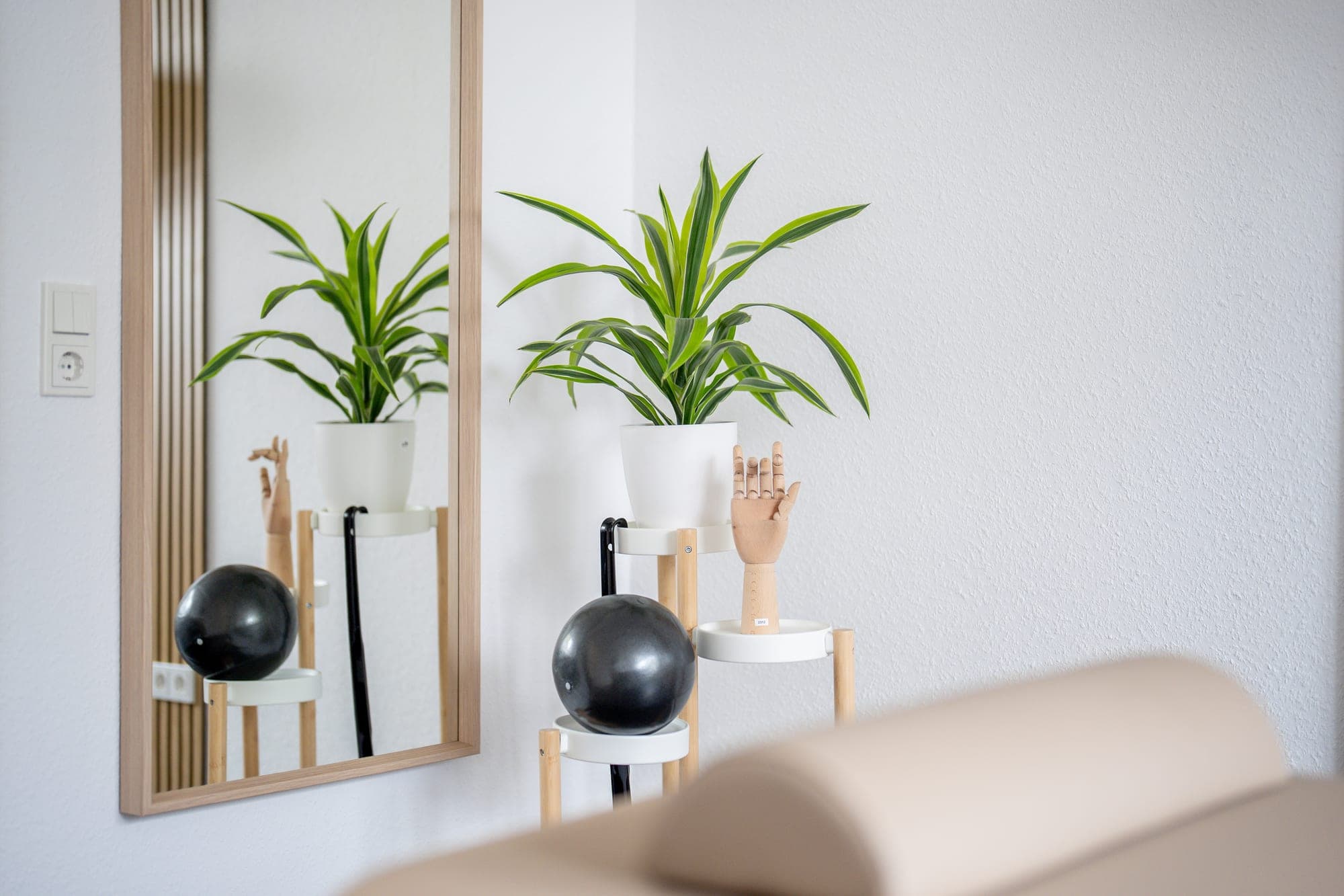 Ein Spiegel hängt an einer weißen Wand neben einem Pflanzenständer mit einer Pflanze, einer Holzhand und einem schwarzen Ball.