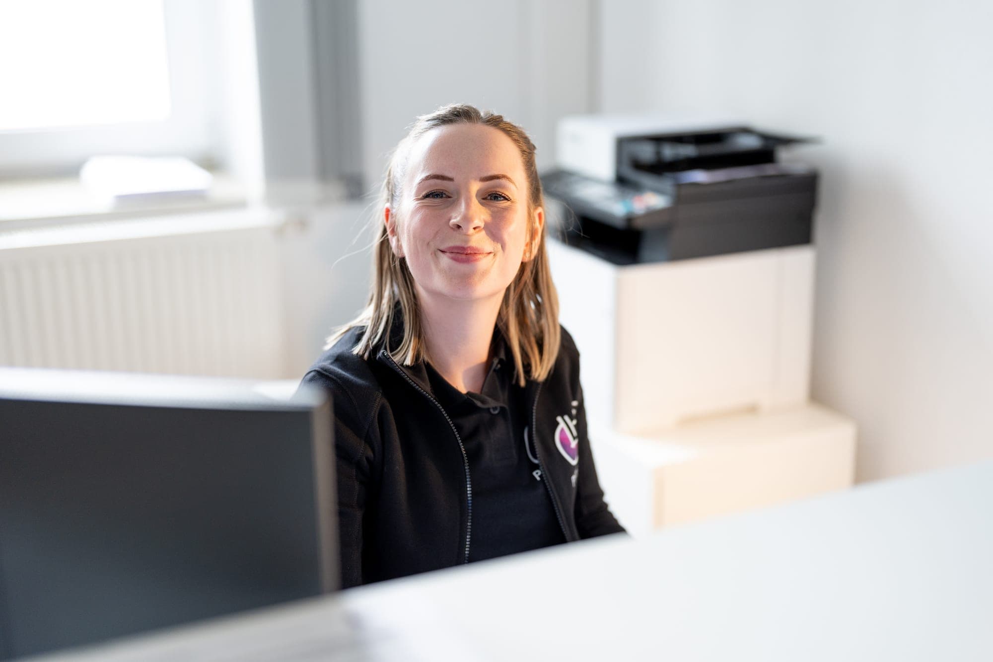 Eine junge Frau mit blondem Haar sitzt an einem Schreibtisch in einem hellen Büro und lächelt in die Kamera.