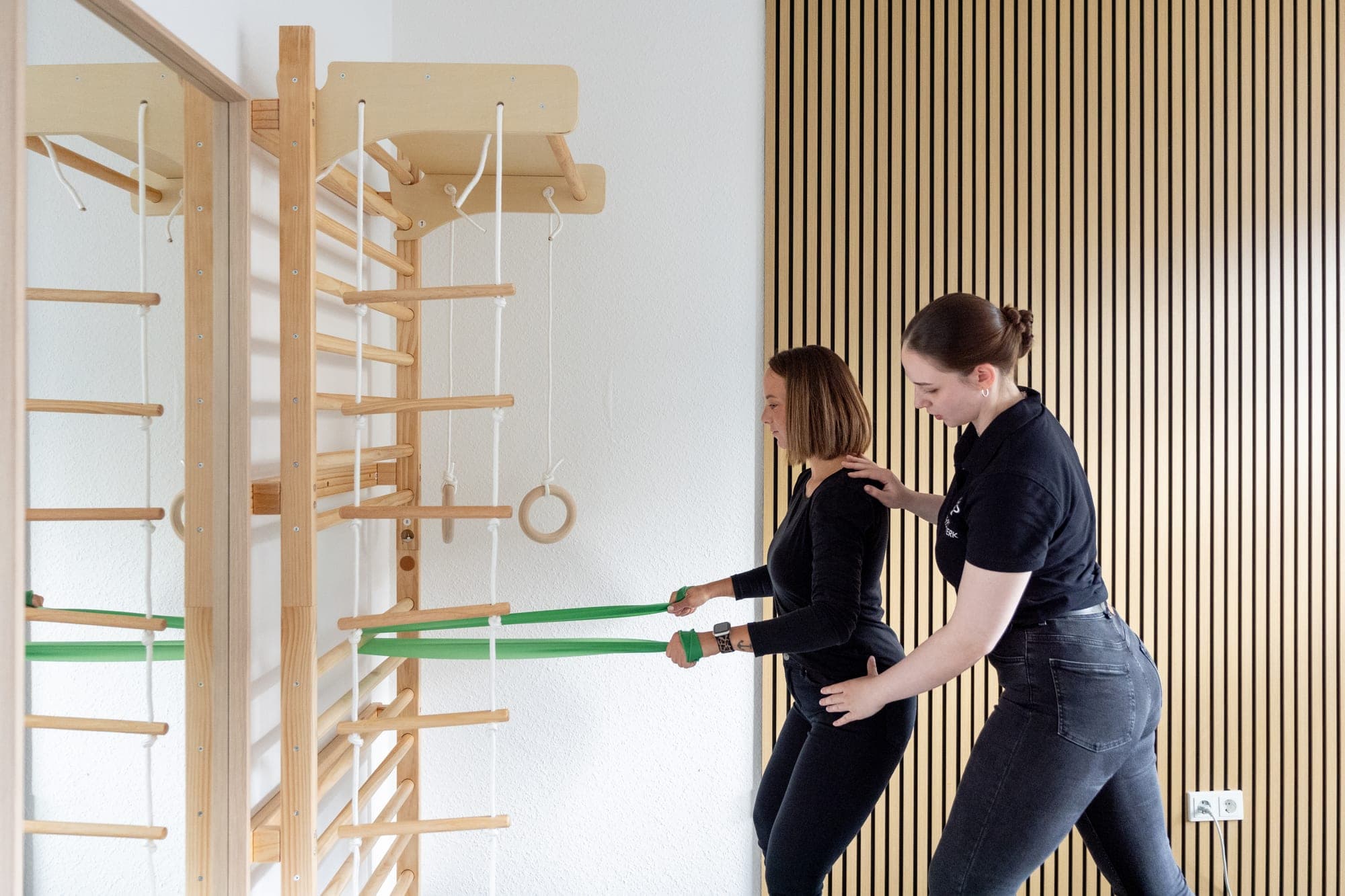 Eine Physiotherapeutin korrigiert die Haltung einer Frau, die mit einem grünen Widerstandsband an einer hölzernen Sprossenwand trainiert.
