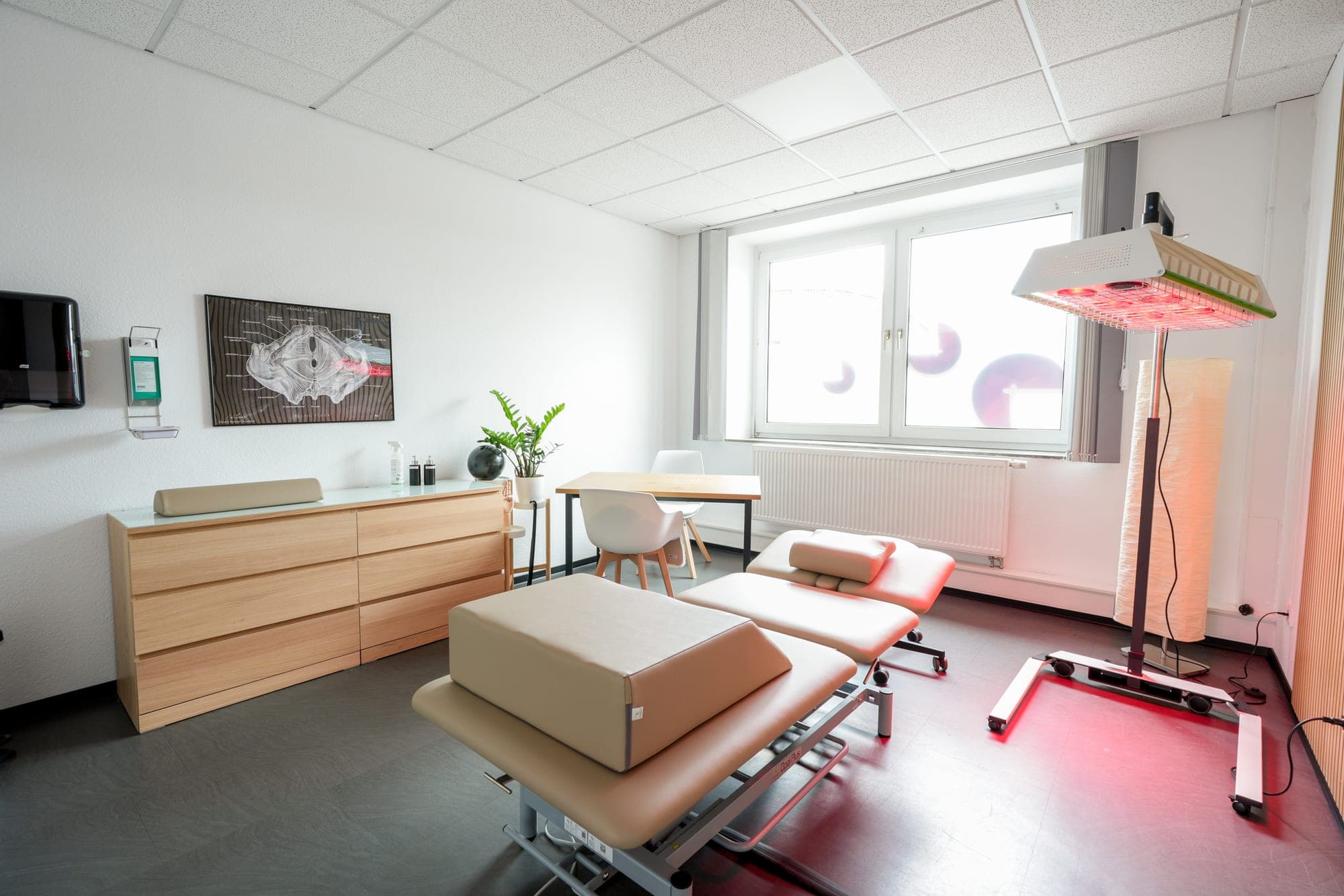 Ein heller Behandlungsraum mit einer Therapieliege, einer Infrarotlampe, einer Holzkommode und einem anatomischen Poster an der Wand.