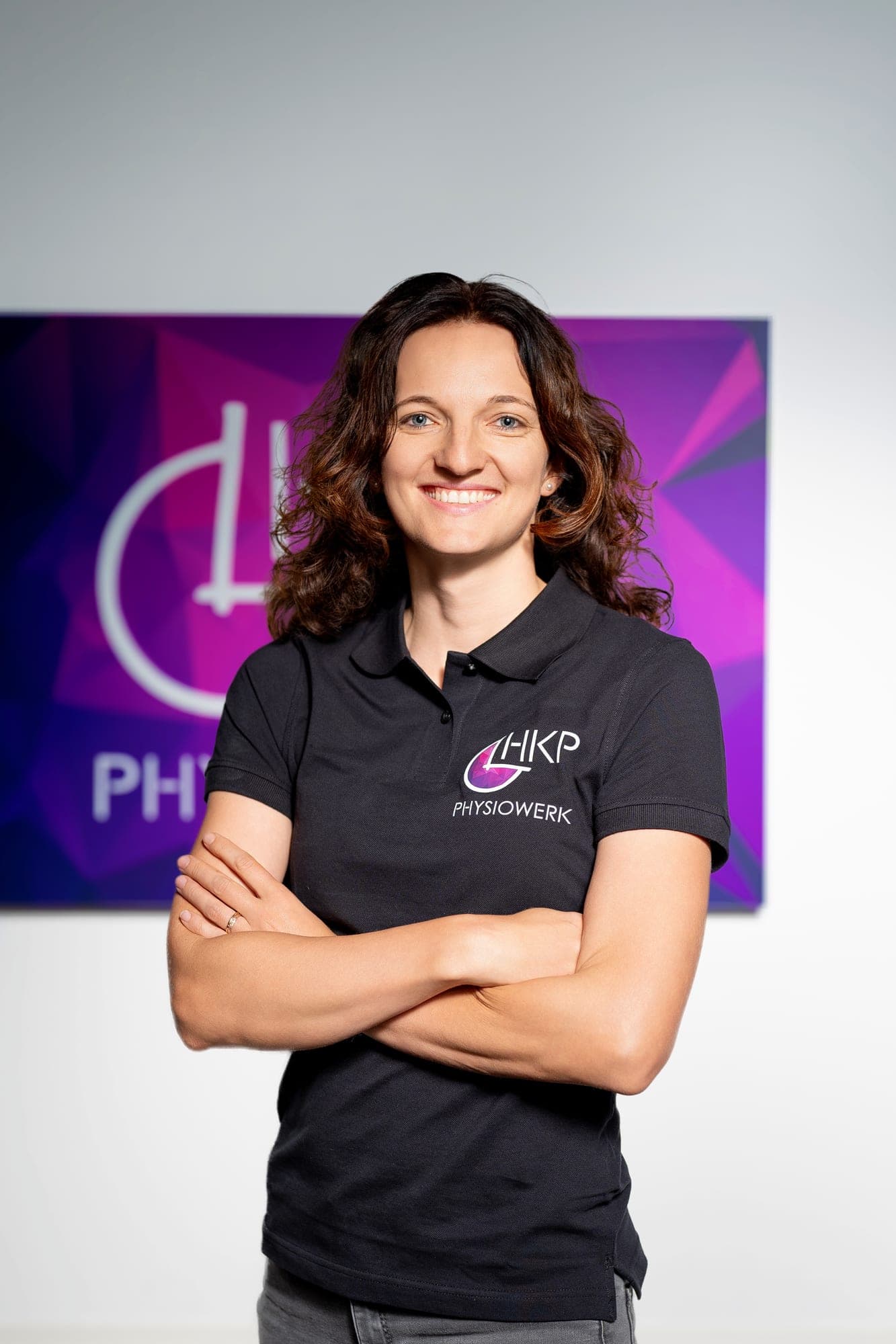 Eine lächelnde Frau mit braunen lockigen Haaren trägt ein schwarzes Polo-Shirt mit Firmenlogo und steht mit verschränkten Armen vor einer violetten Wand mit einem weißen Logo.