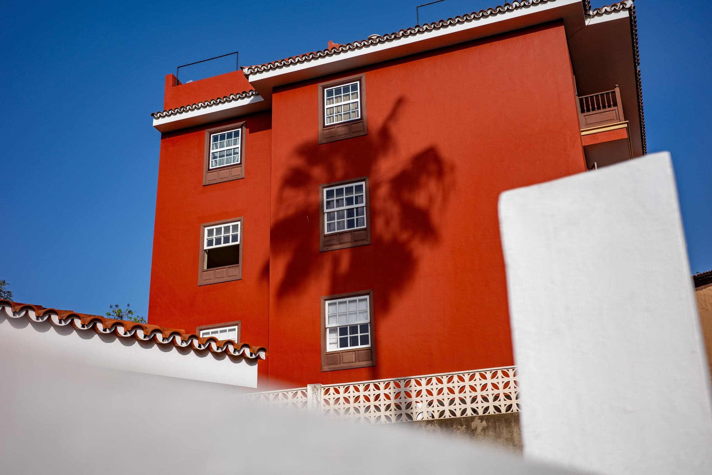 Eine leuchtend rote Hausfassade mit mehreren Fenstern und einem deutlichen Schatten einer Palme, davor eine weiße Mauer, unter einem klaren blauen Himmel.
