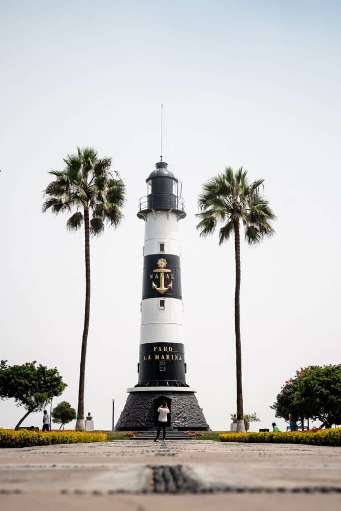 Ein schwarz-weißer Leuchtturm mit einem goldenen Ankersymbol steht zentral zwischen zwei hohen Palmen in einem Park.