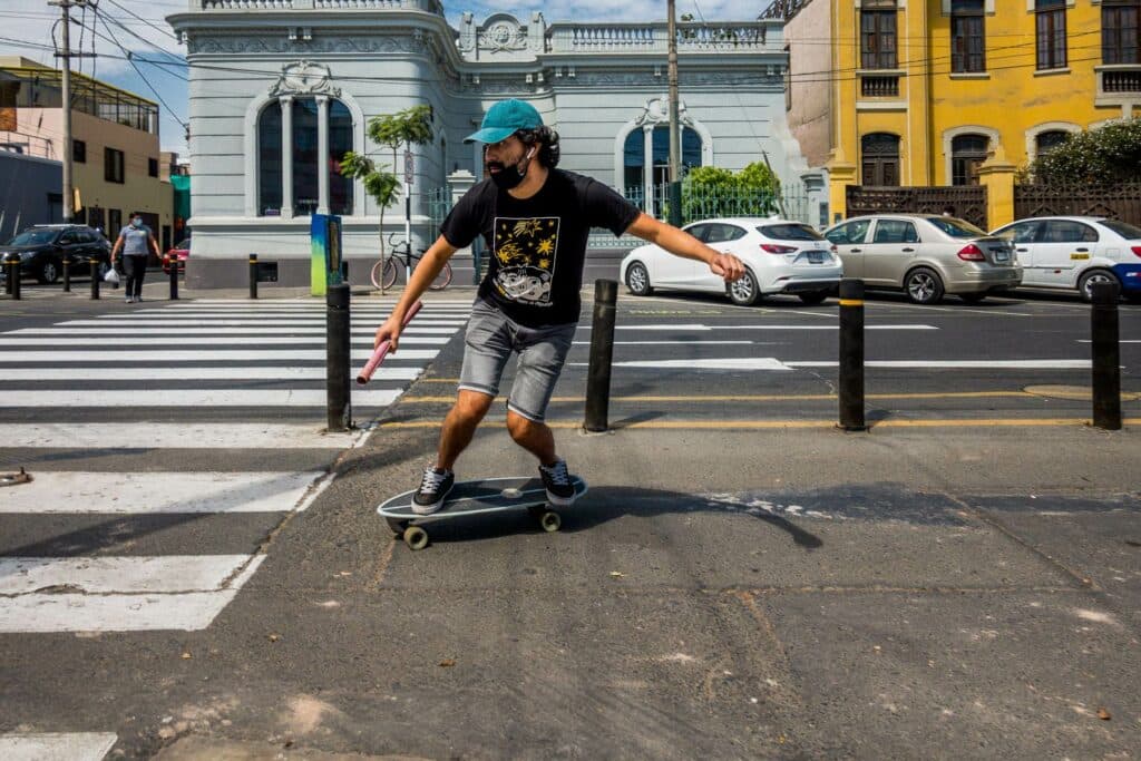 Ein junger Mann mit Cap und schwarzem T-Shirt fährt auf einem Skateboard über einen Zebrastreifen vor historischen Gebäuden.