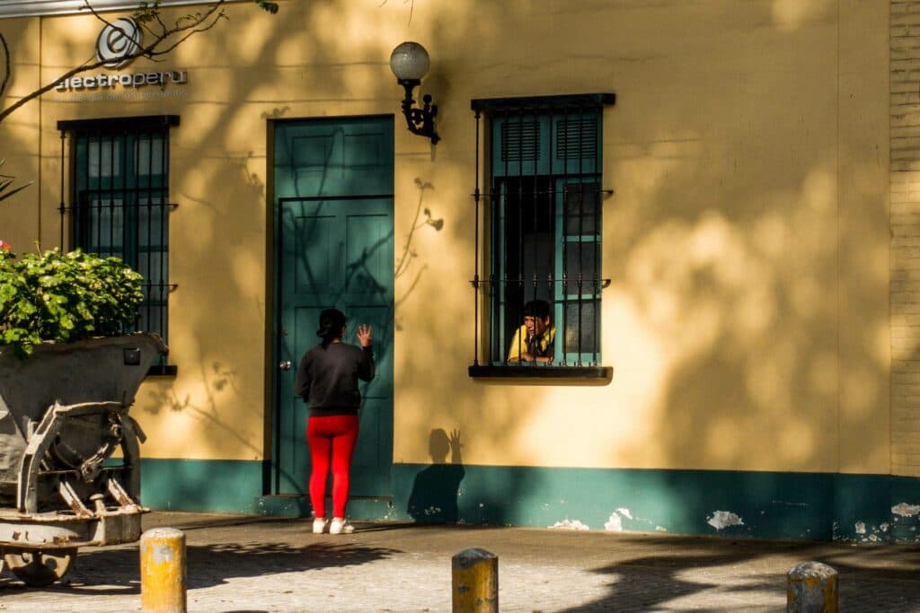 Eine Person in roter Hose steht vor einer gelben Hausfassade und winkt einer anderen Person zu, die aus einem vergitterten Fenster schaut.