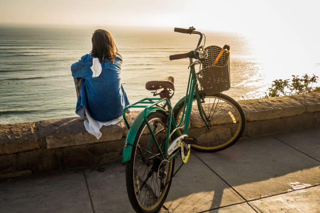 Eine Person mit dunklen Haaren sitzt in einer blauen Jacke auf einer steinernen Mauer und blickt auf das glitzernde Meer, daneben steht ein grünes Fahrrad mit einem Korb.