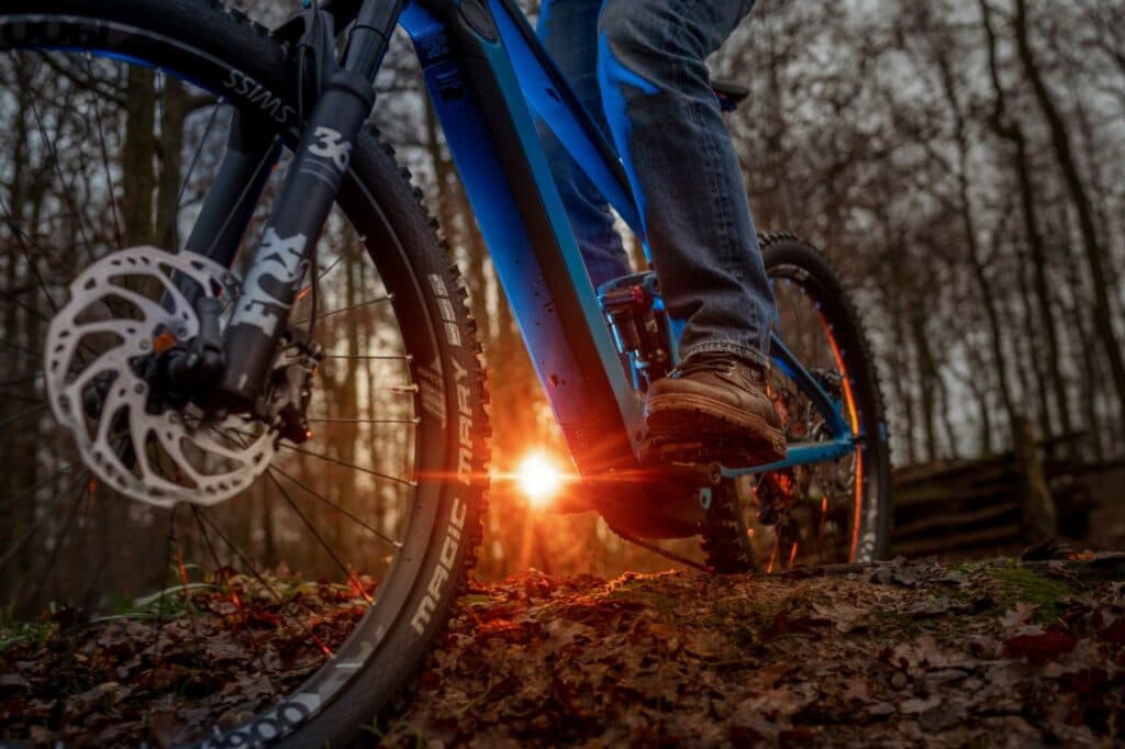Nahaufnahme eines blauen Mountainbikes auf einem Waldboden, wobei die tiefstehende Sonne einen hellen Lichtpunkt erzeugt.