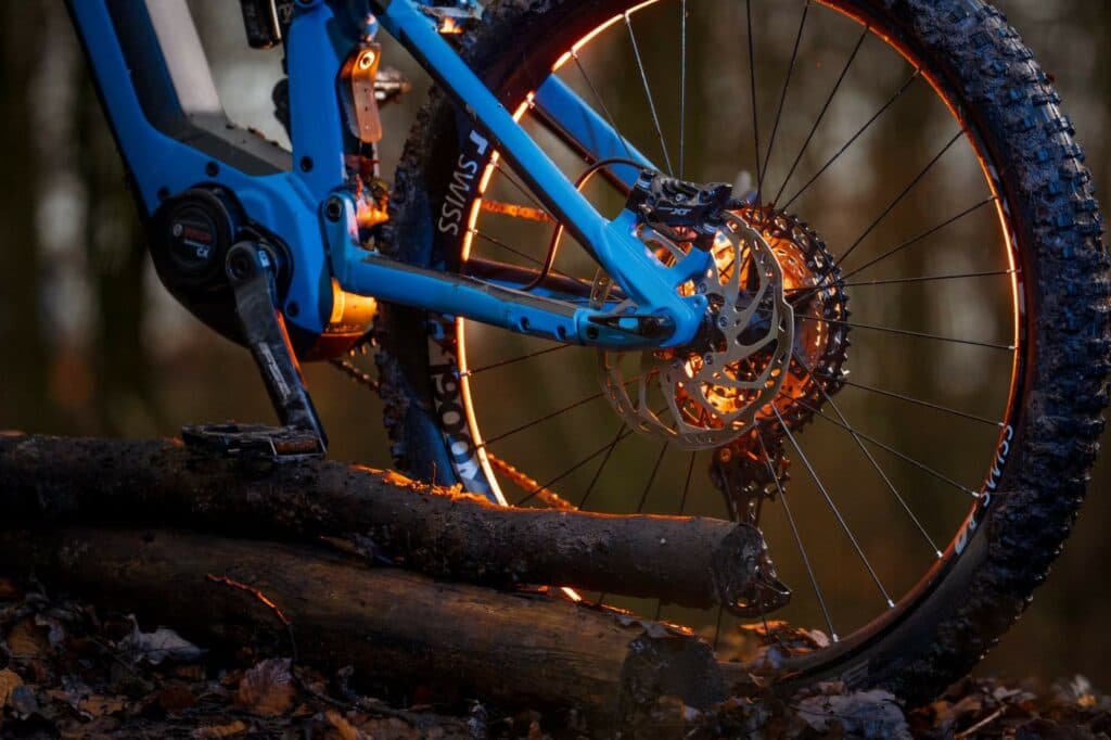 Detailansicht des Hinterrads und Rahmens eines blauen Mountainbikes, das im Gegenlicht der untergehenden Sonne steht und dessen Kanten golden leuchten.