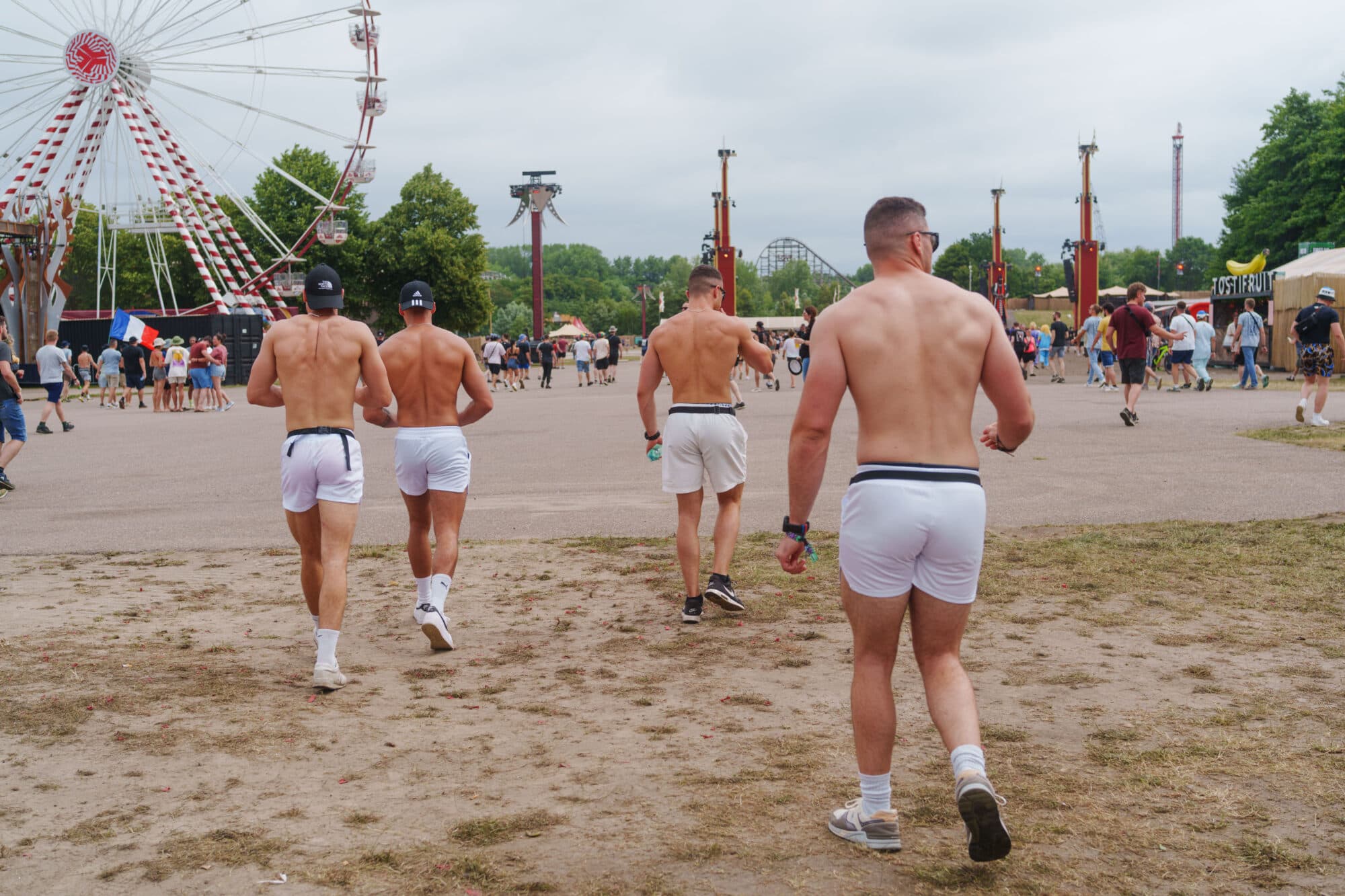 Vier muskulöse Männer mit freiem Oberkörper und weißen Shorts gehen von der Kamera weg über einen Platz auf einem Volksfest, im Hintergrund sind ein Riesenrad und weitere Fahrgeschäfte zu sehen.