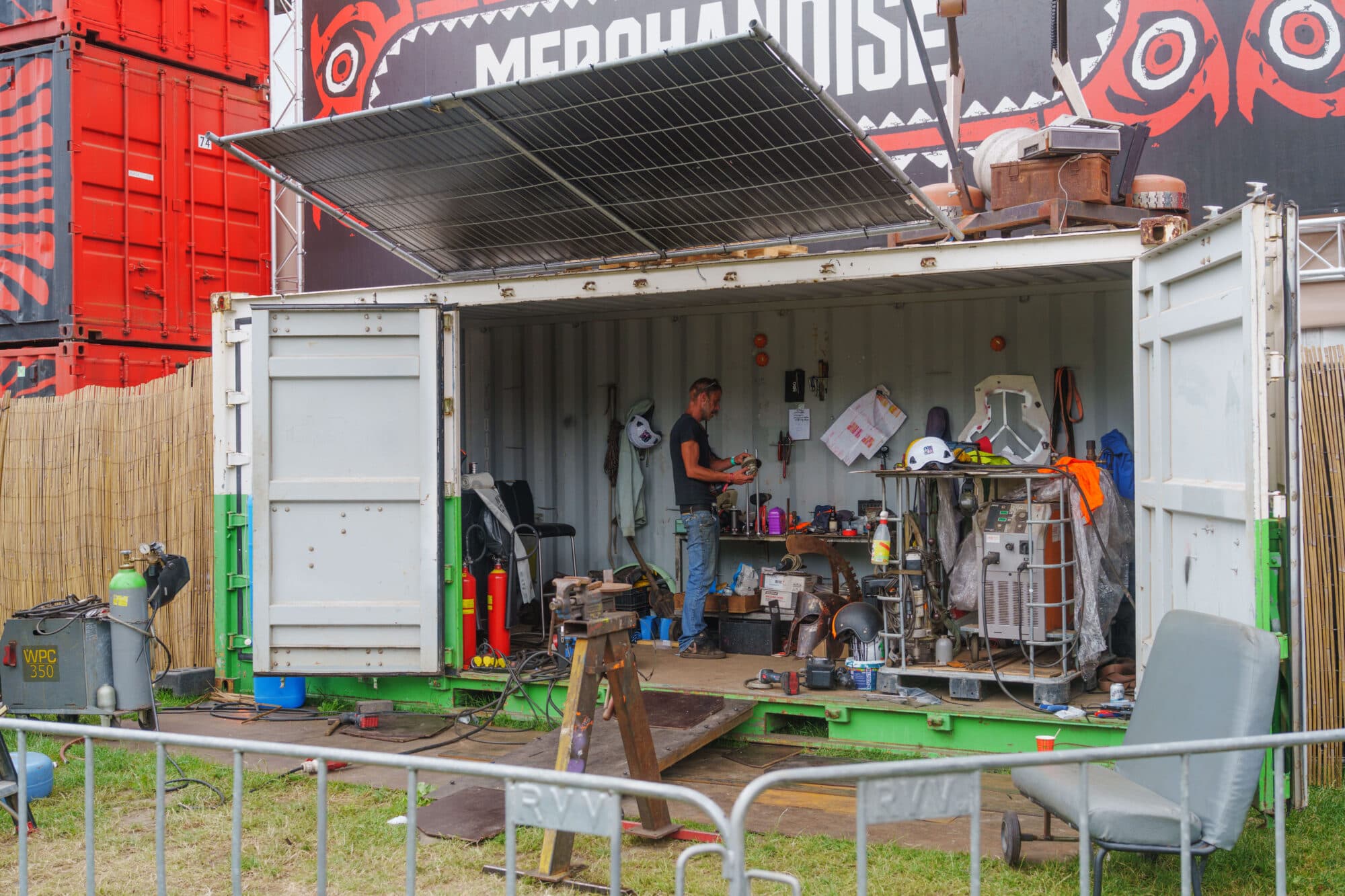Ein Mann steht in einem geöffneten Schiffscontainer, der als Werkstatt dient, und arbeitet an einem Tisch voller Werkzeuge.