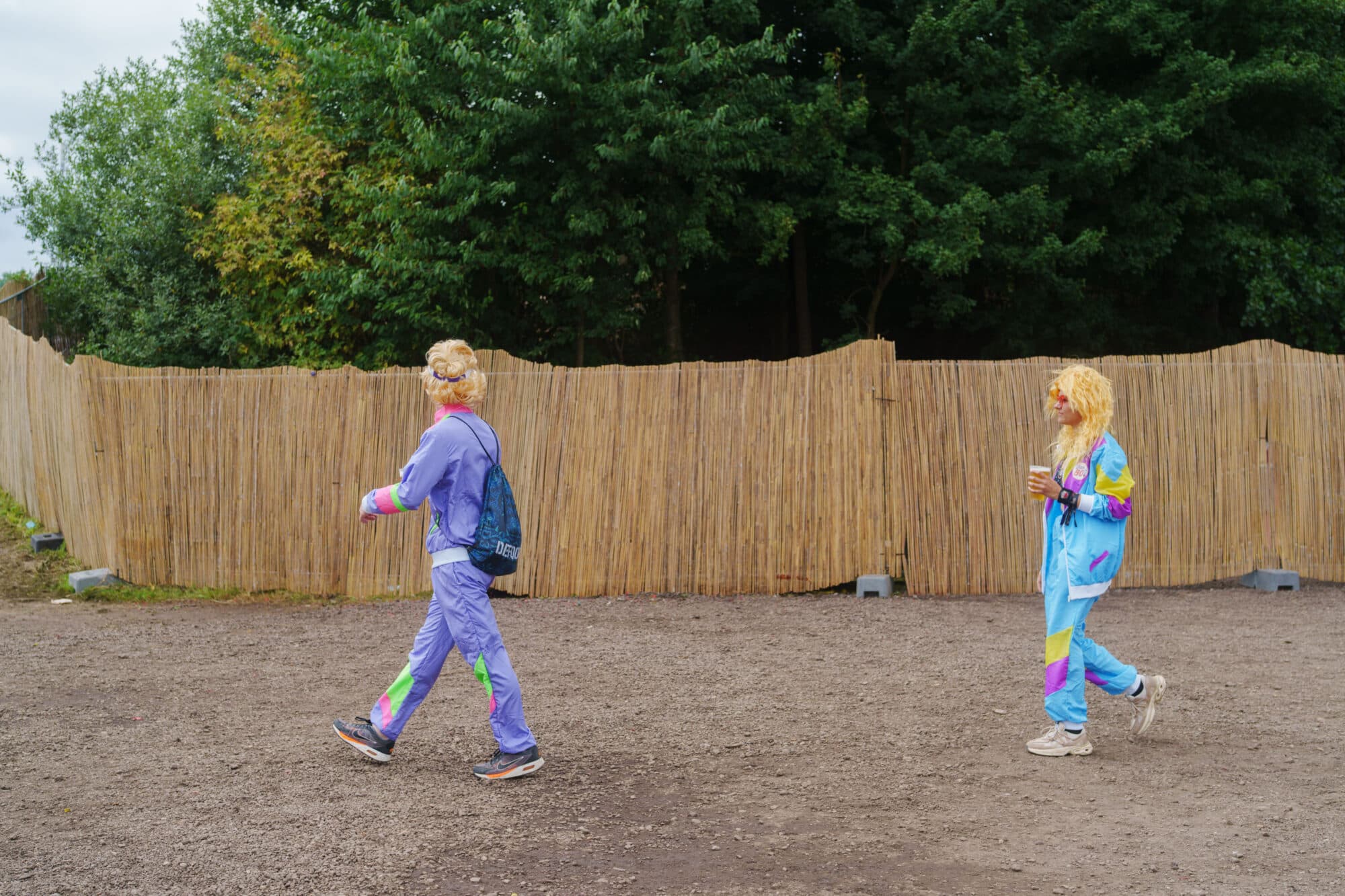 Zwei blondierte Personen in bunter Sportkleidung im Retro-Stil gehen auf einem unbefestigten Weg vor einem Schilfzaun und Bäumen.