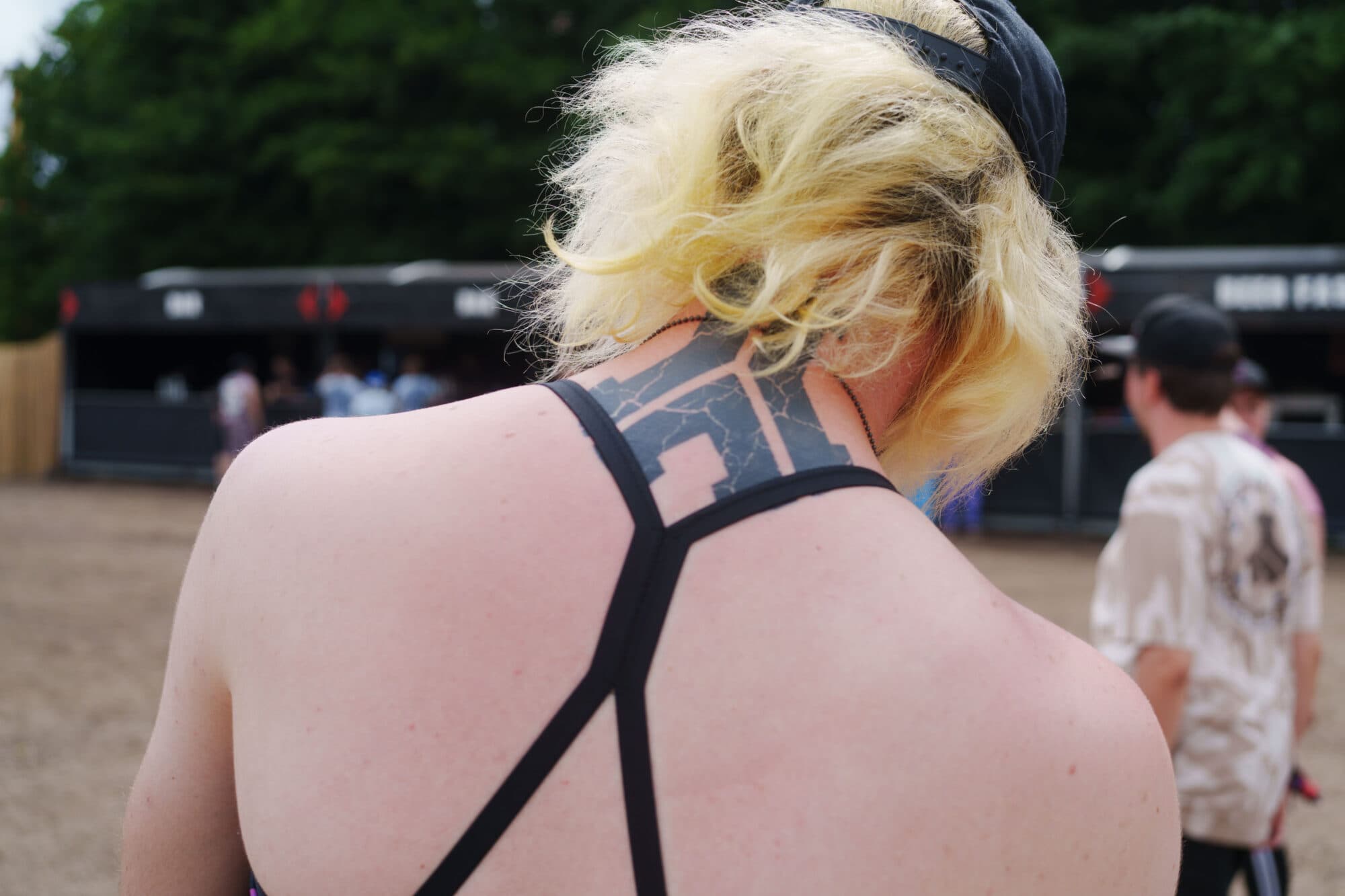 Nahaufnahme von hinten auf den Nacken und die Schultern einer Person mit blonden Haaren und einem geometrischen schwarzen Tattoo im Nacken, bekleidet mit einem schwarzen Trägertop.