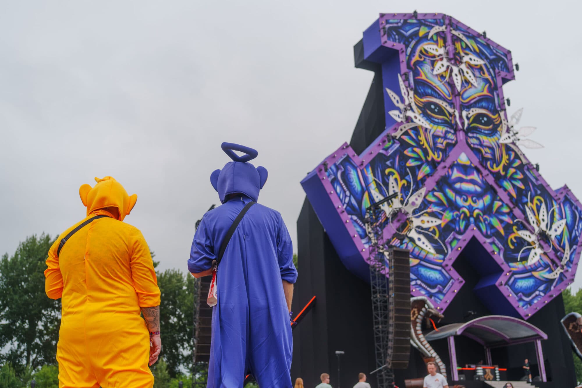 Zwei Personen in gelben und lila Ganzkörperkostümen stehen mit dem Rücken zur Kamera vor einer riesigen, aufwendig gestalteten Festivalbühne.