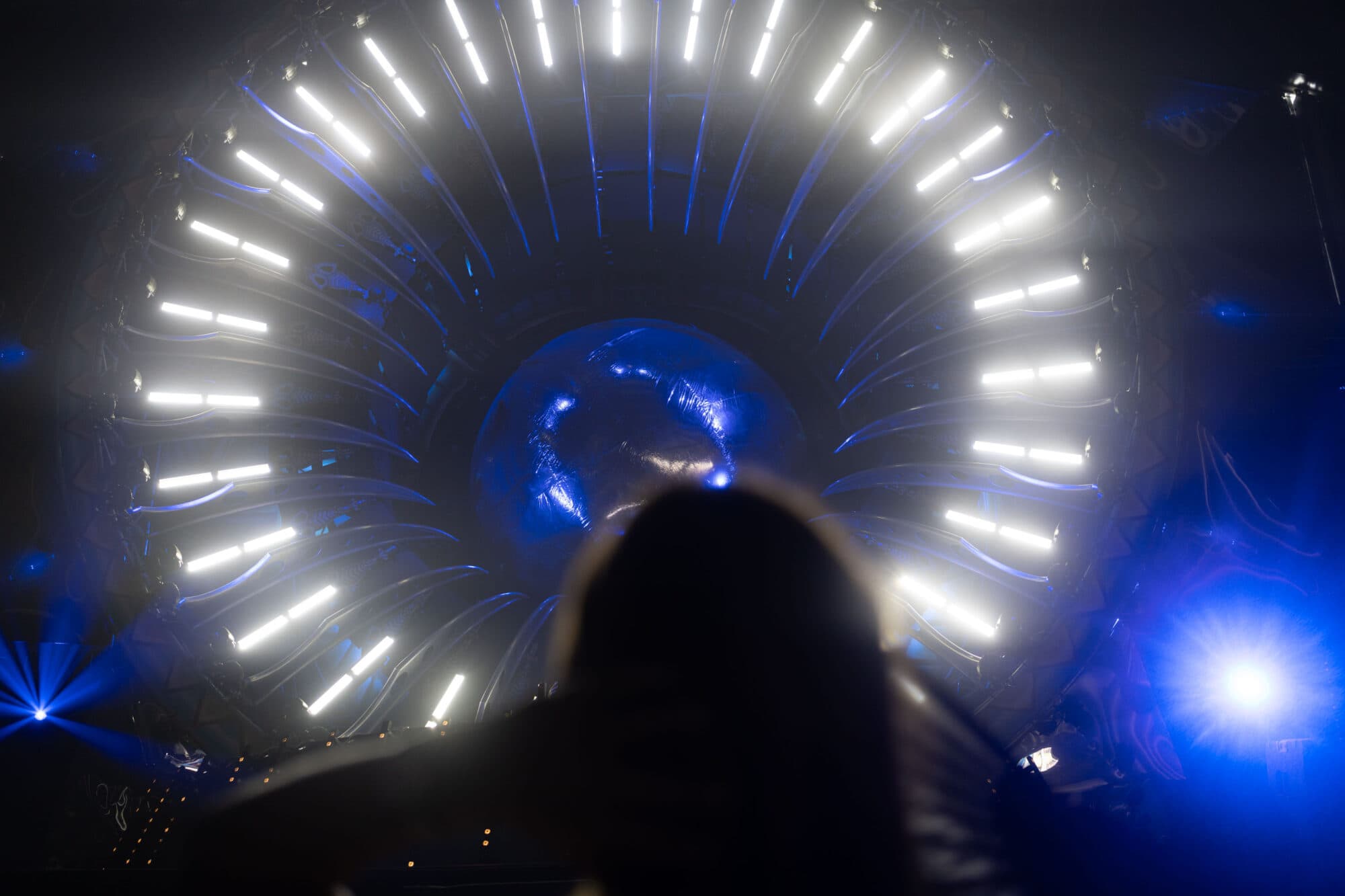 Eine unscharfe Silhouette einer Person im Vordergrund vor einer hell leuchtenden kreisförmigen Lichtinstallation in einem dunklen Raum.