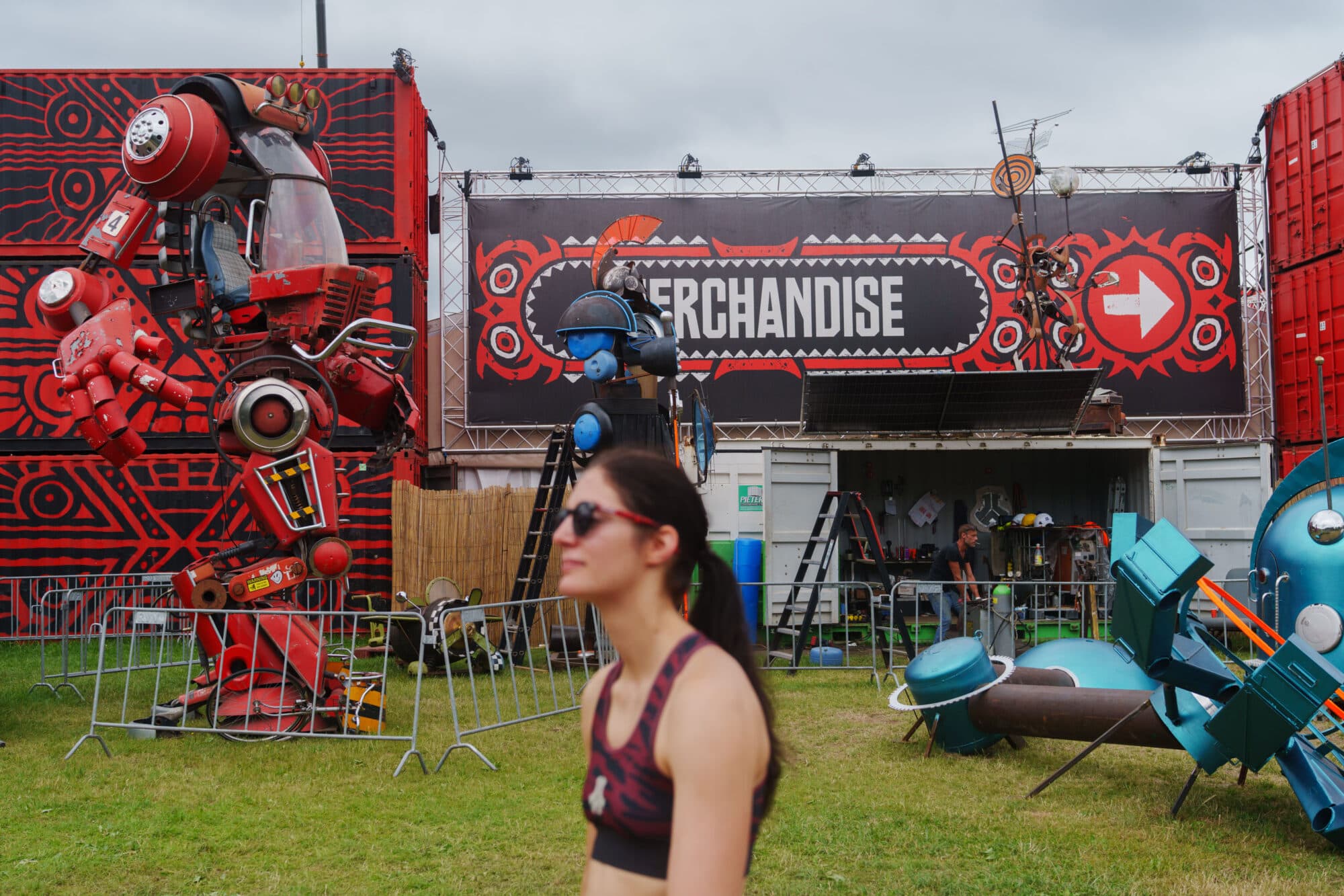 Eine junge Frau mit Sonnenbrille steht im Vordergrund vor einem Merchandise-Stand auf einem Festivalgelände mit großen roten Roboterskulpturen.