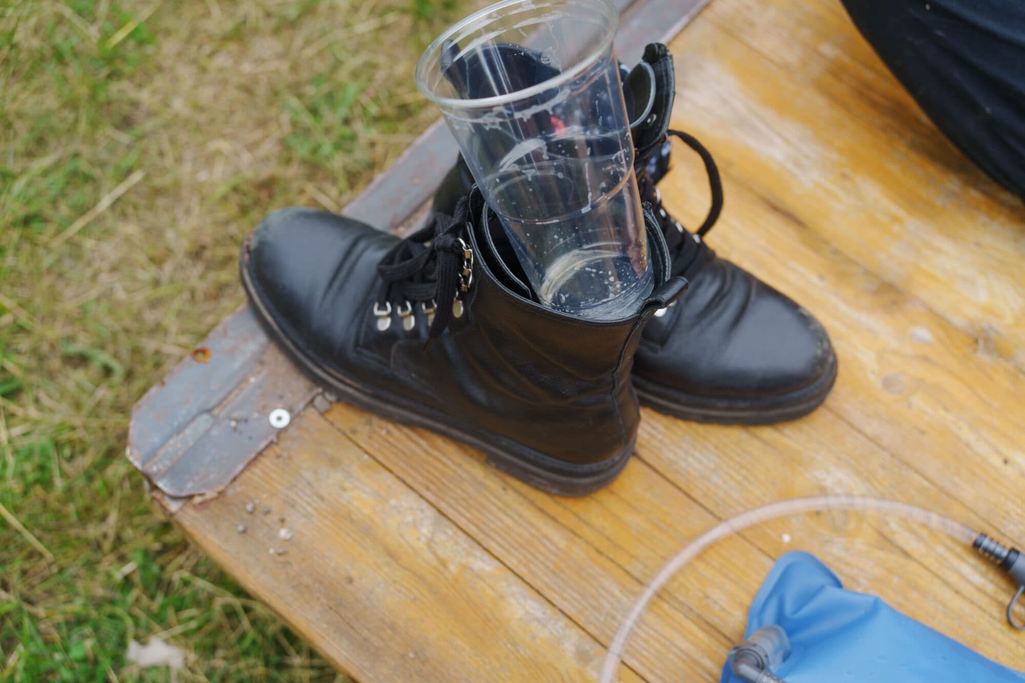 Ein schwarzer Stiefel steht auf einer hölzernen Unterlage, in seinem Schaft steckt ein leerer, durchsichtiger Plastikbecher.