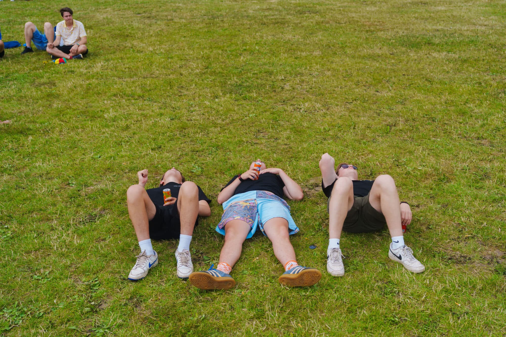 Drei junge Männer liegen nebeneinander auf dem Rücken auf einer grünen Wiese und schauen in den Himmel, ein weiterer Mann sitzt im Hintergrund.