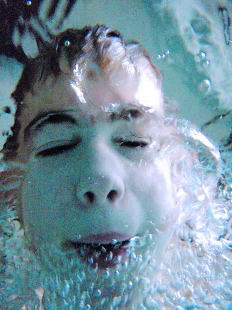 Ein Gesicht unter Wasser mit geschlossenen Augen, umgeben von zahlreichen aufsteigenden Luftblasen.