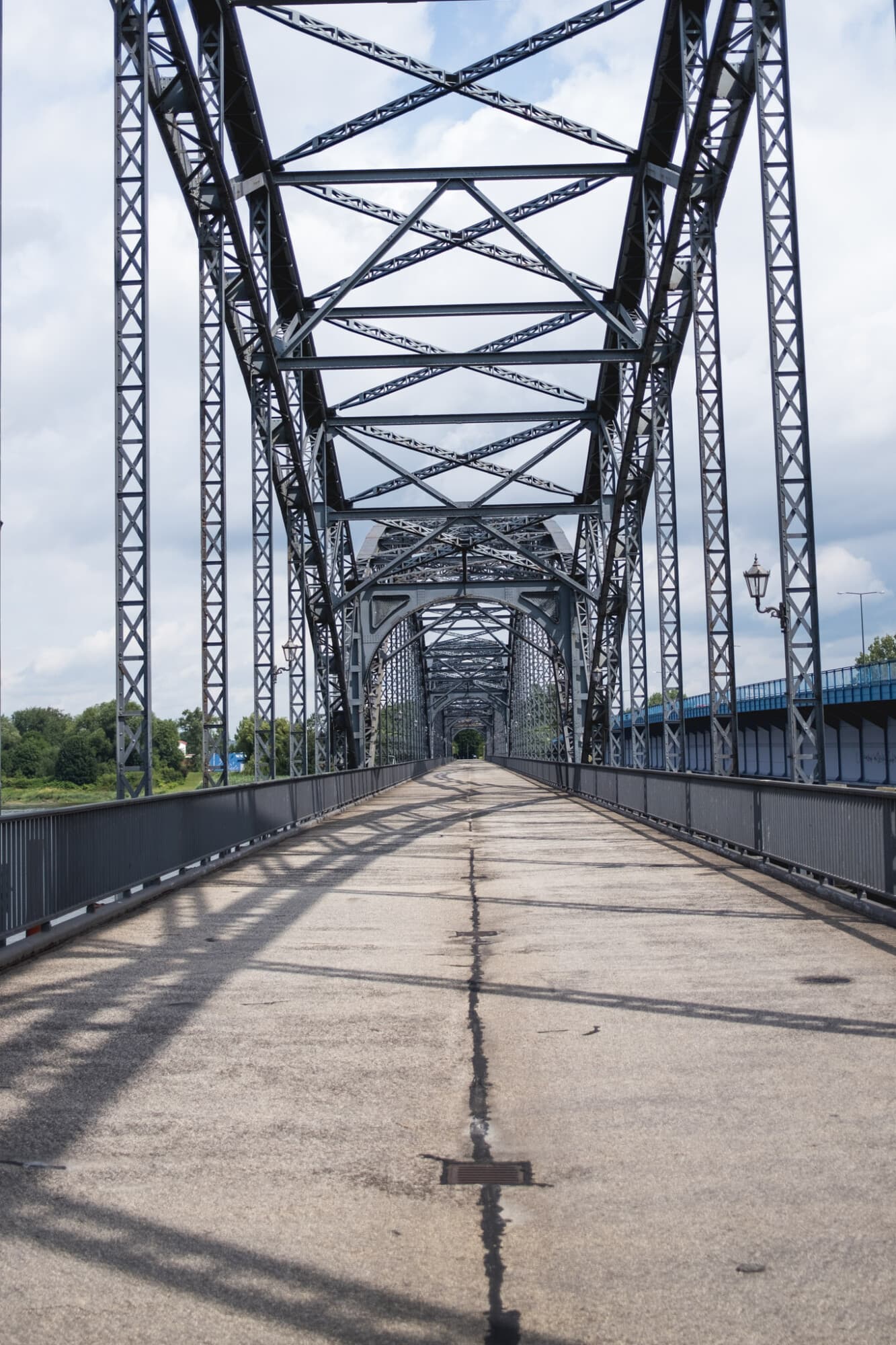 Eine leere, asphaltierte Brücke führt zentral in die Tiefe, überdacht von einer komplexen, grauen Stahlfachwerkkonstruktion unter einem bewölkten Himmel.