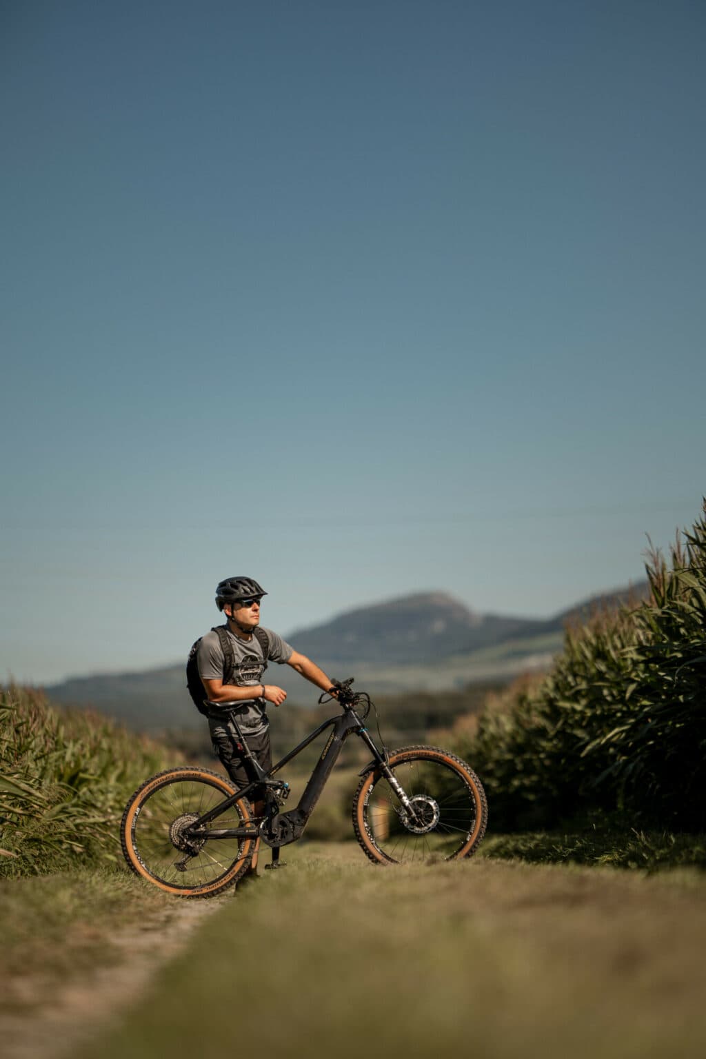 Ein Mann mit Helm und Sportkleidung steht auf einem Feldweg mit seinem Mountainbike und blickt in die Ferne vor einer hügeligen Landschaft.