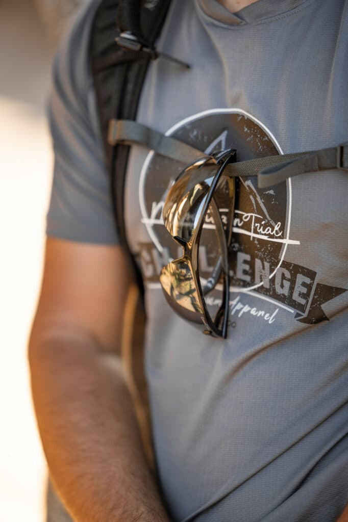 Nahaufnahme des Oberkörpers einer Person in grauem Sport-T-Shirt mit einem Rucksack, an dessen Schulterriemen eine Sonnenbrille befestigt ist.