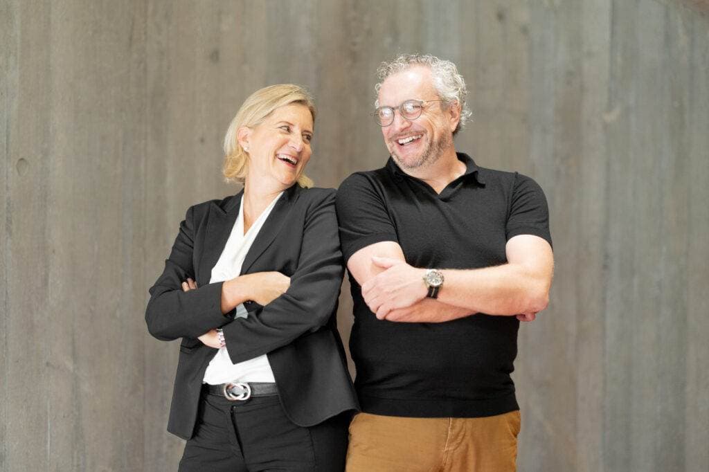 Ein Mann und eine Frau stehen nebeneinander vor einer grauen Betonwand, beide haben die Arme verschränkt und lachen sich an.