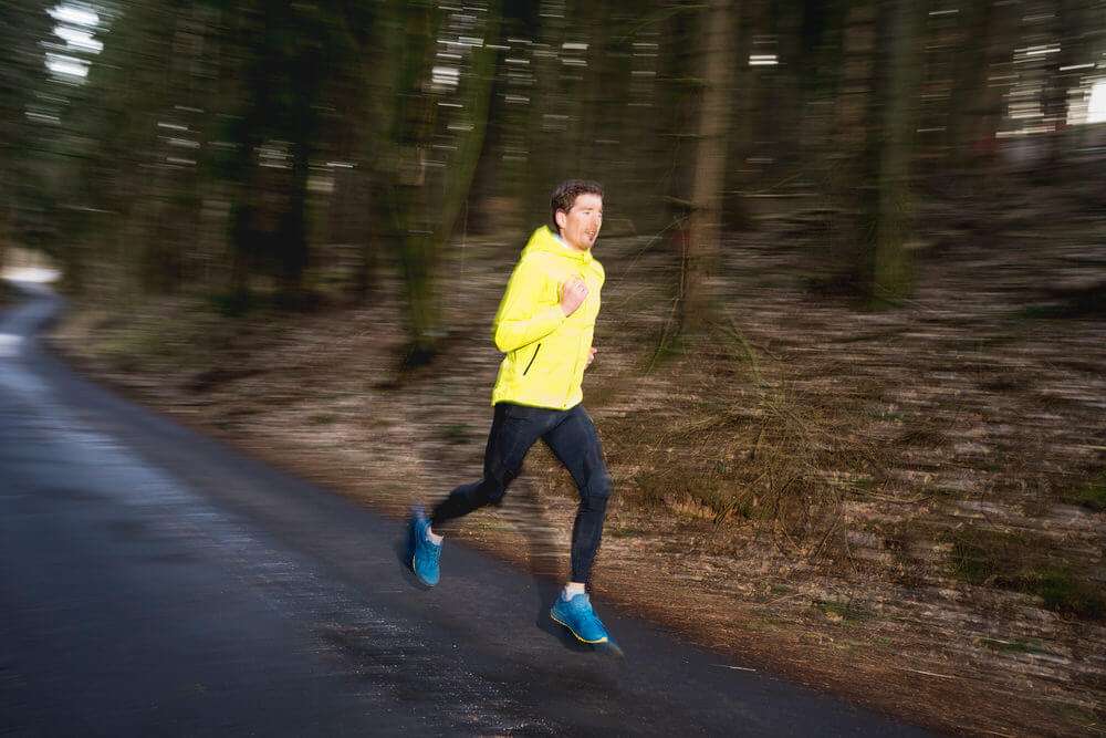 Ein Mann in einer leuchtend gelben Sportjacke und dunkler Hose läuft auf einer asphaltierten Straße durch einen Wald, wobei der Hintergrund durch eine Mitzieher-Technik verwischt ist.