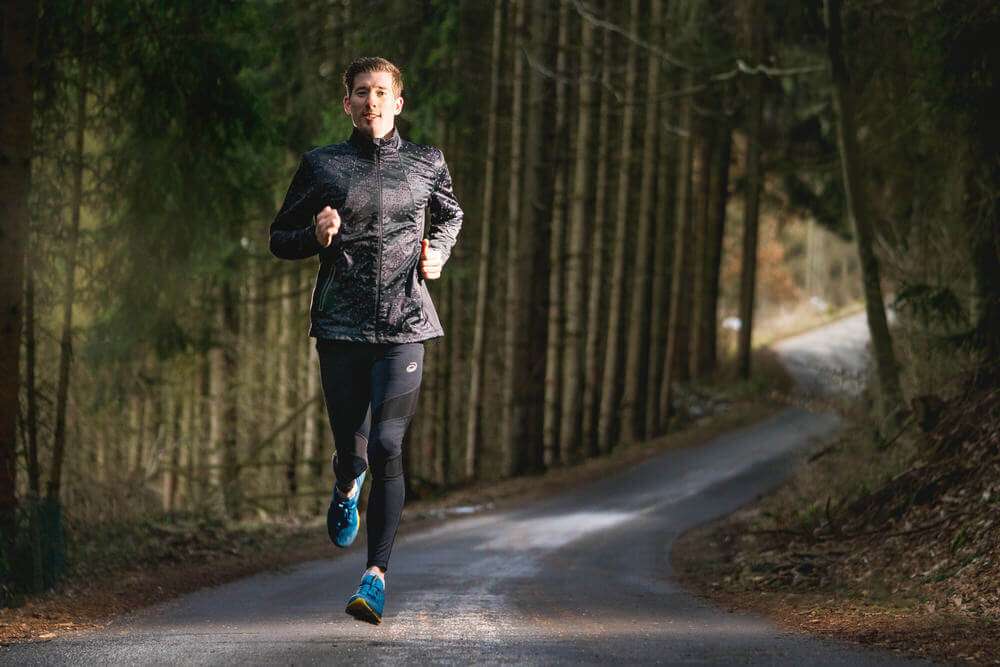 Ein junger Mann in schwarzer Sportkleidung läuft auf einer asphaltierten Straße durch einen Wald.