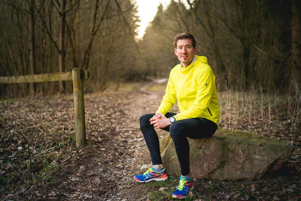 Ein Mann in einer neongelben Sportjacke und schwarzen Leggings sitzt auf einem großen Stein an einem Waldweg und blickt in die Kamera.